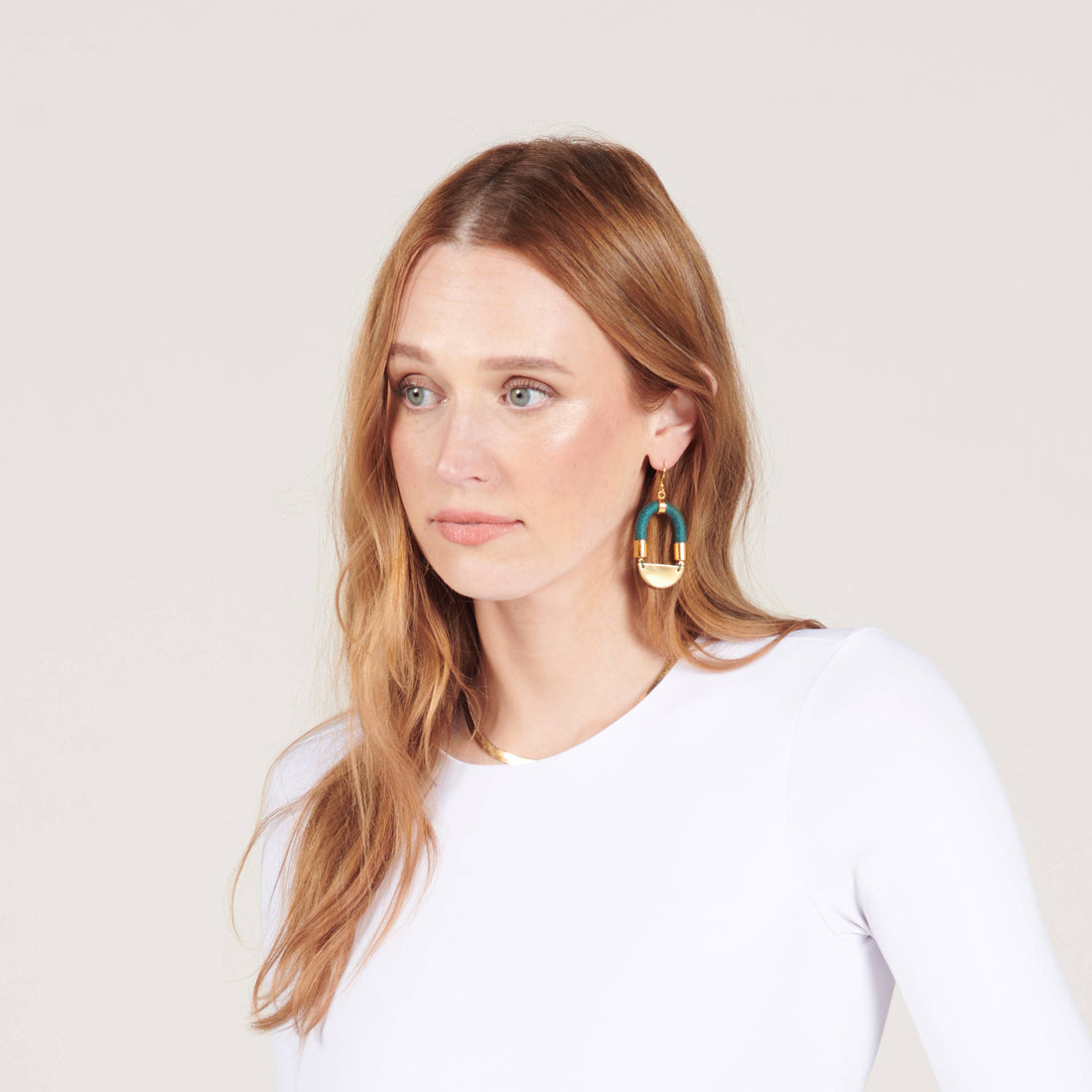 Half Moon earrings, teal yarn-wrapped cording with gold-toned brass, worn on model's ear, looking to the side.