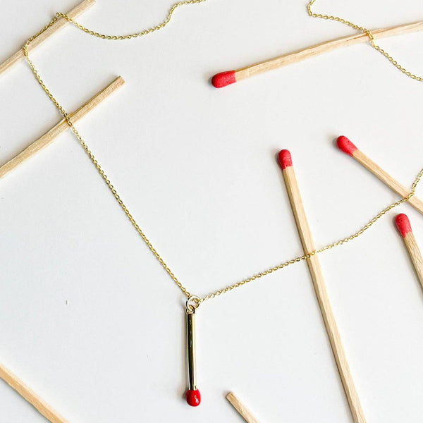 Natalie Clare Matchstick Necklace, gold-plated with red tip pendant, flat lay surrounded by real matchsticks.