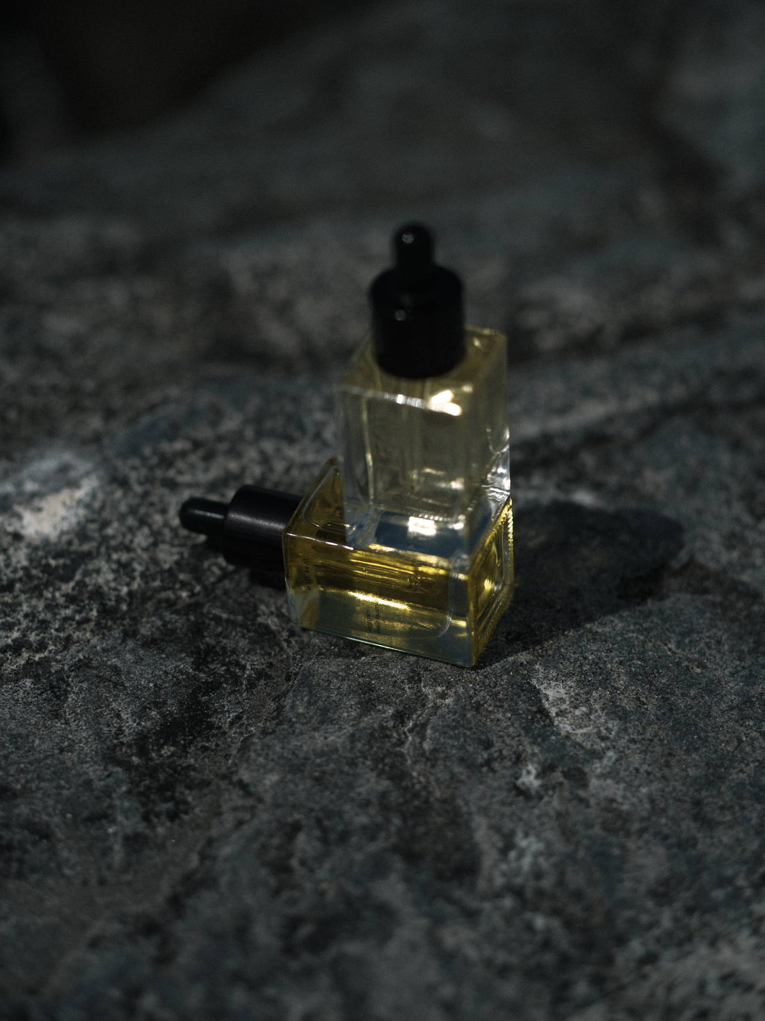 Shave Oil, two clear glass dropper bottles with golden yellow oil, one standing, one lying, close-up on dark textured stone.