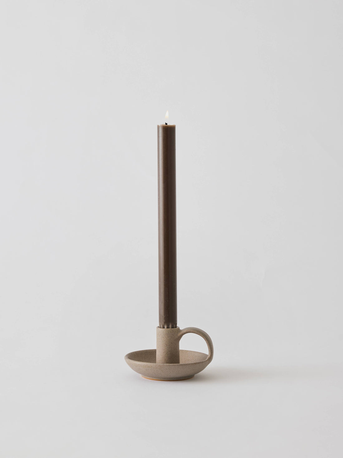 Nimo candle holder, matte speckled stoneware with side handle, holding a lit brown taper candle, front view.