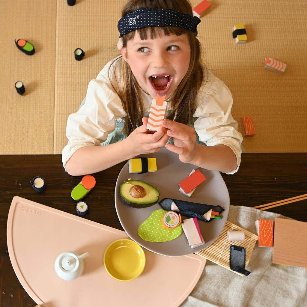 Smiling child holds a striped wooden sushi toy from Sushi Party Set to mouth, with colorful beechwood and fabric play food…