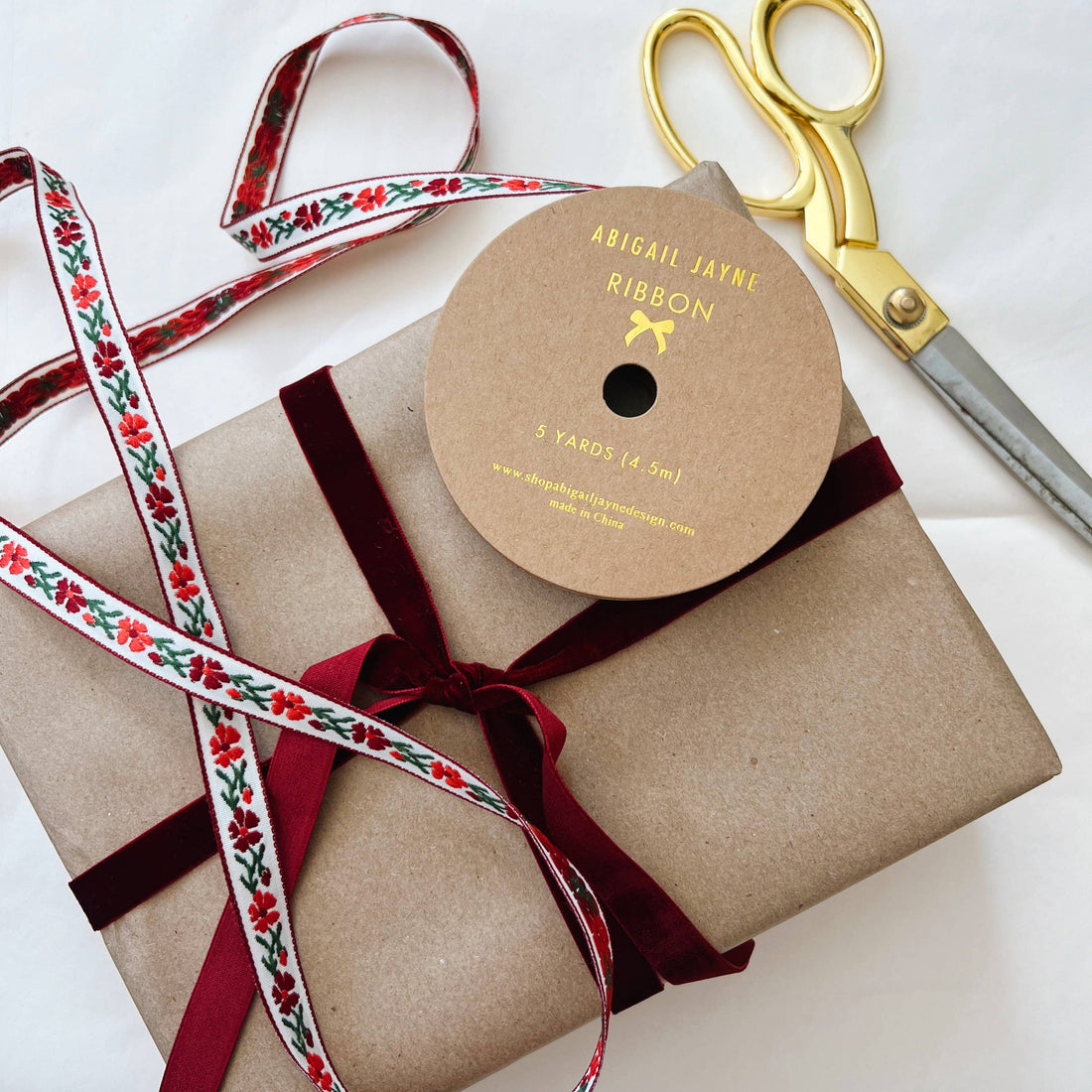 Garnet velvet ribbon spool beside a gift wrapped with matching velvet ribbon and a white floral ribbon, with gold scissors.