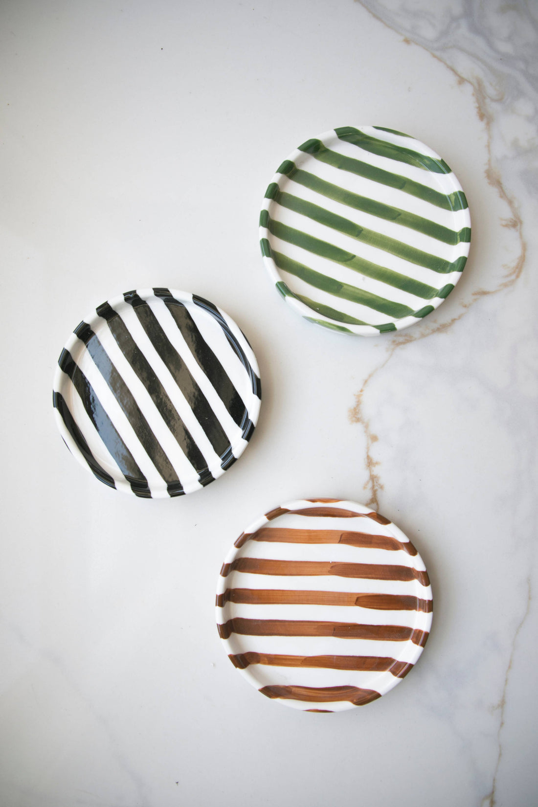 Striped Ceramic Plates, white ceramic trio with black, green, and brown hand-painted stripes, flat lay on marble surface.