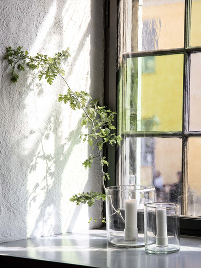 
                    
                      Luna clear recycled glass candle holders, a pair with lit white candles on a sunlit sill, reflecting colored window light.
                    
                  