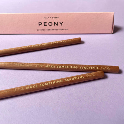 
                    
                      Imogen Owen Peony Scented Pencils, three cedarwood writing pencils with gold foil lettering, angled in front of pink box.
                    
                  