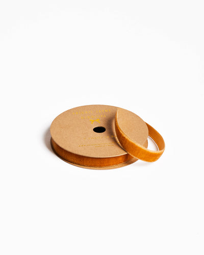 
                    
                      Amber velvet ribbon on kraft paper spool, partially unwound, showing product label, angled view.
                    
                  