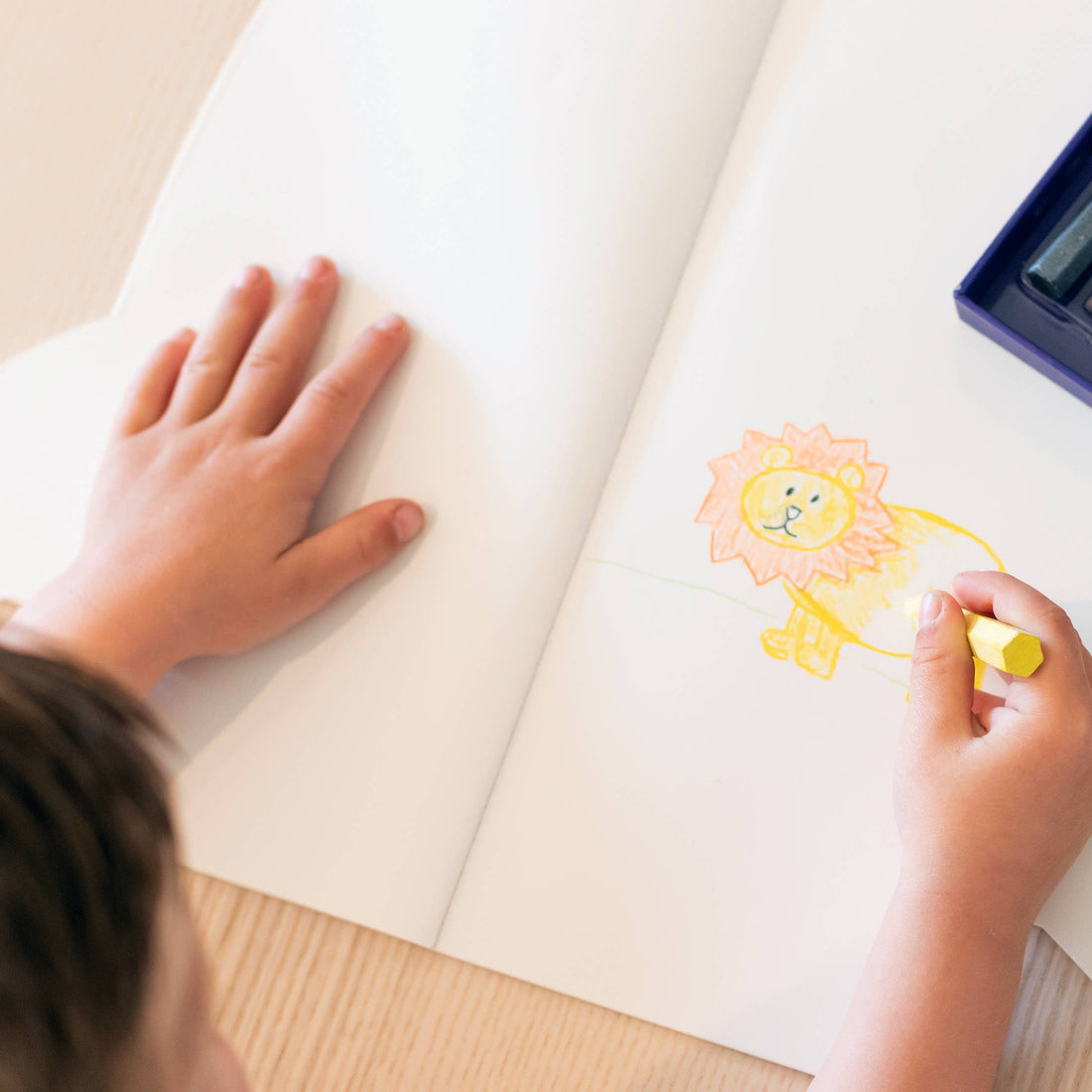 Jumbo Rocket Paper Coloring Book open to a lion drawing, child's hands coloring with a yellow crayon, top-down view.
