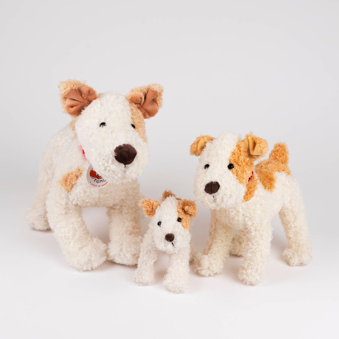 Eliot Stuffed Dog, cream and brown plush with red collar and label, standing with two smaller stuffed dogs, showing scale.