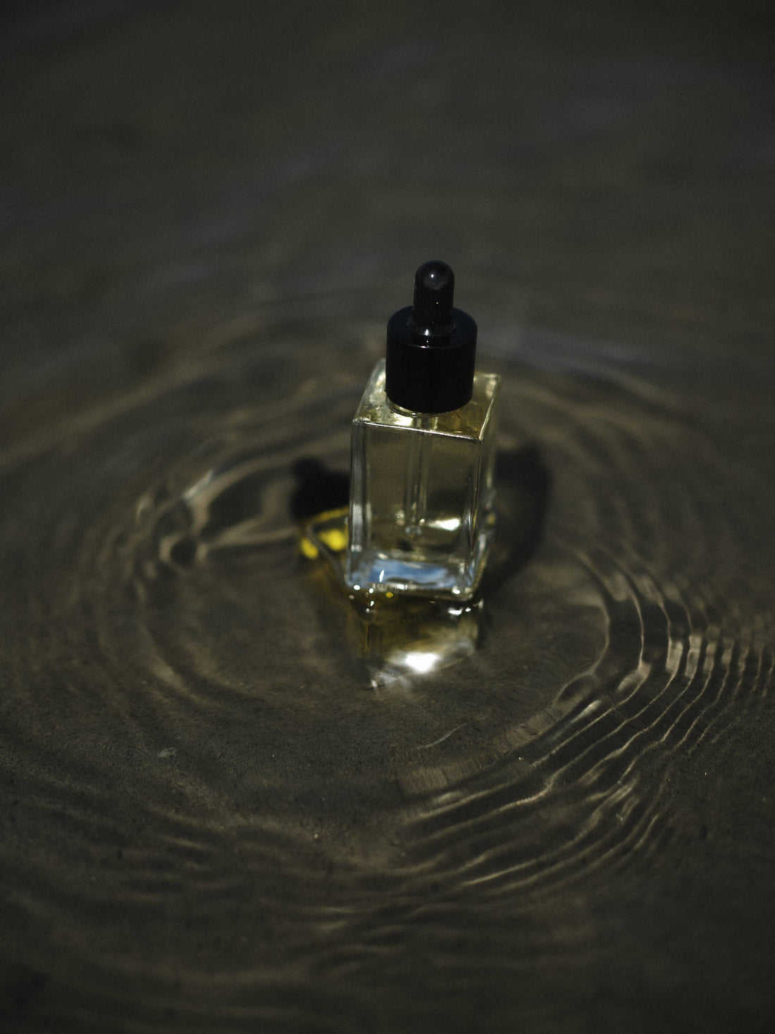 Shave Oil in square clear glass bottle with light golden oil and black dropper, resting in dark rippling water.