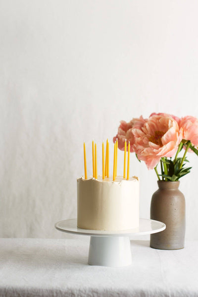 
                    
                      Traditional Beeswax Birthday Candles, yellow pure beeswax, standing in a white frosted cake, side view with pink peonies.
                    
                  