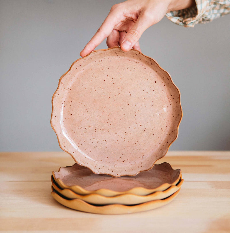 Ohyoko Ceramics Handcrafted Stoneware Plates, speckled blush pink, wavy scalloped edges, one held by hand above a stack of…