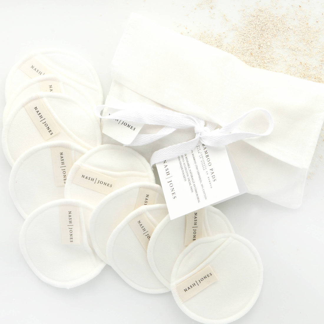 Multiple creamy white Nash and Jones Bamboo and Cotton Reusable Facial Pads, fanned around a matching drawstring bag with…