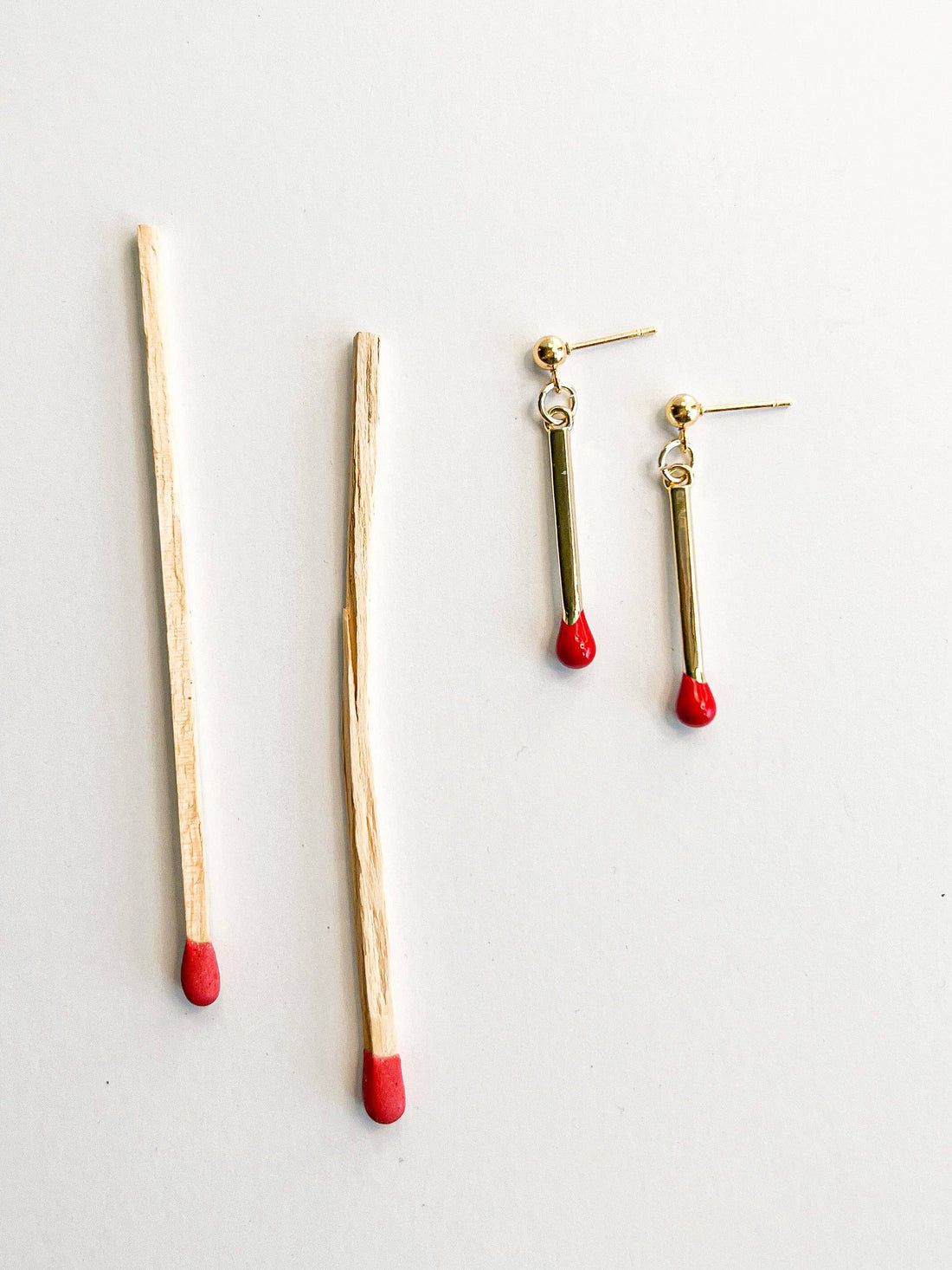 Matchstick drop earrings, 18k gold plated with red tips, flat lay next to real matchsticks for scale comparison.