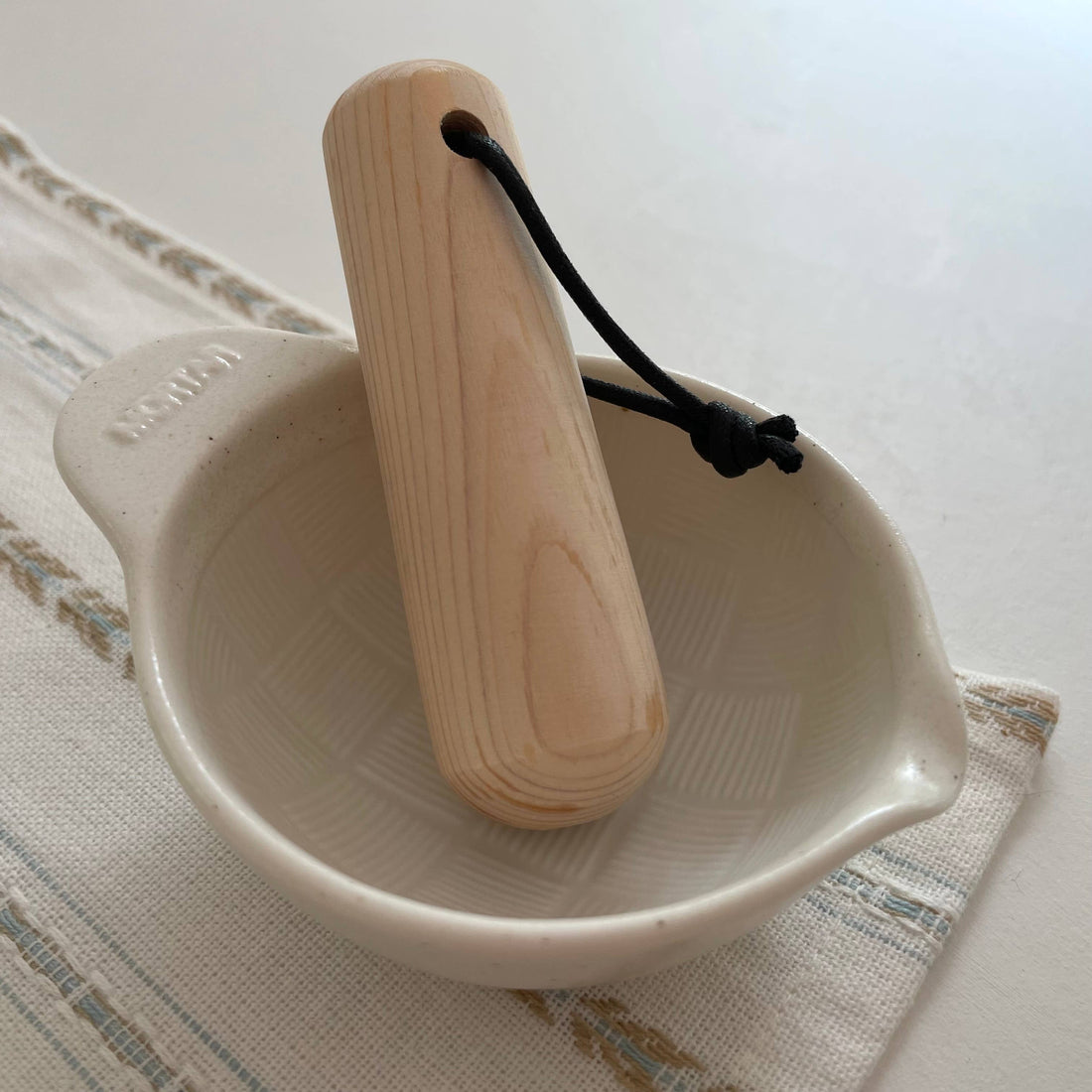 Japanese Ceramic Mortar, white porcelain with textured interior, natural wood pestle with black cord, angled top-down view.