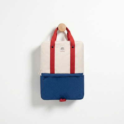 
                    
                      Lefrik Daily 15" Bauhaus Block backpack, beige and royal blue Oxford RPET fabric with red handles, hanging on wooden peg.
                    
                  