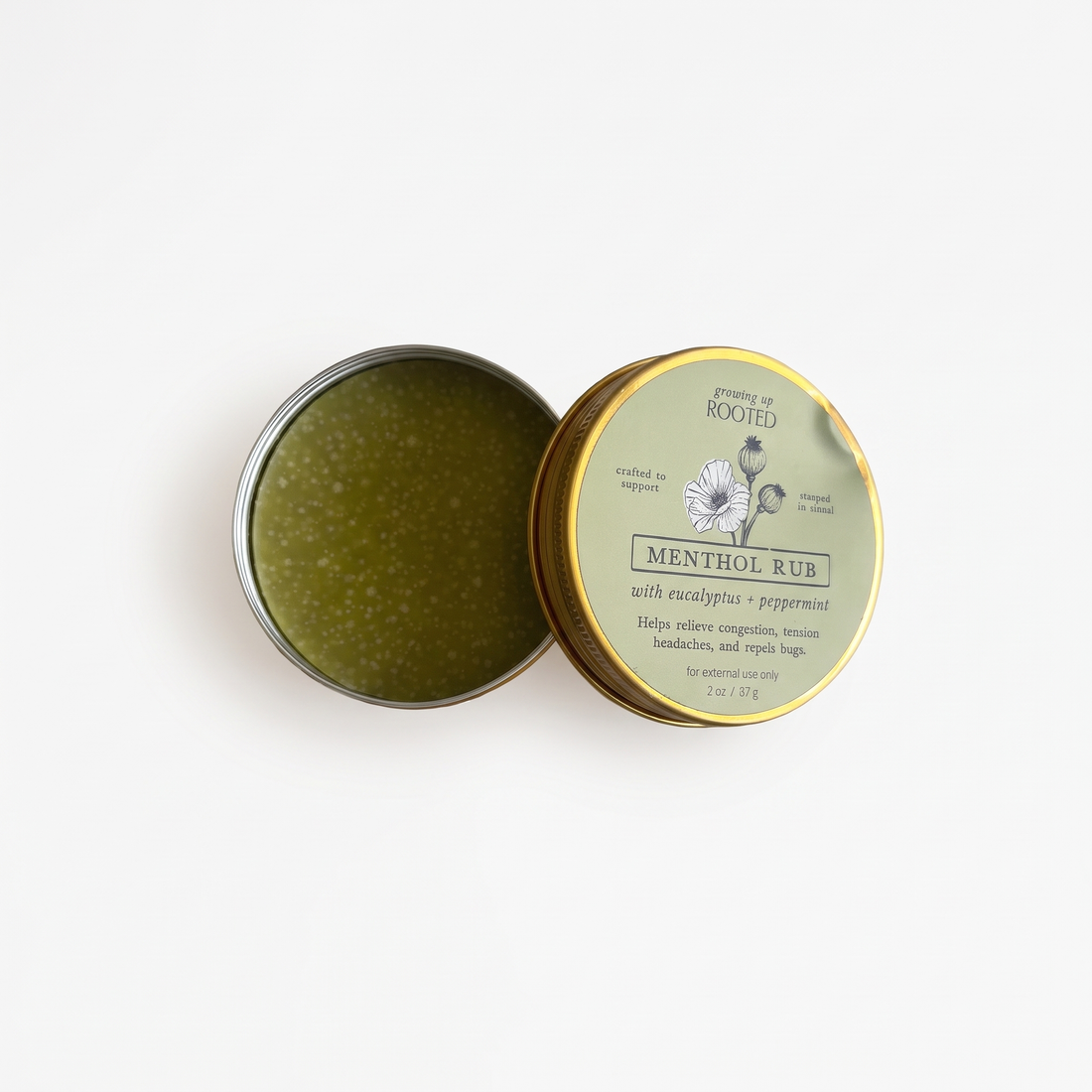 Growing Up Rooted Menthol Rub, open tin showing green rub with white specks, gold-rimmed sage green label, flat lay.