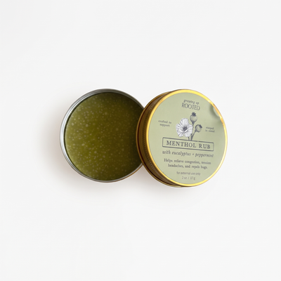 
                    
                      Growing Up Rooted Menthol Rub, open tin showing green rub with white specks, gold-rimmed sage green label, flat lay.
                    
                  