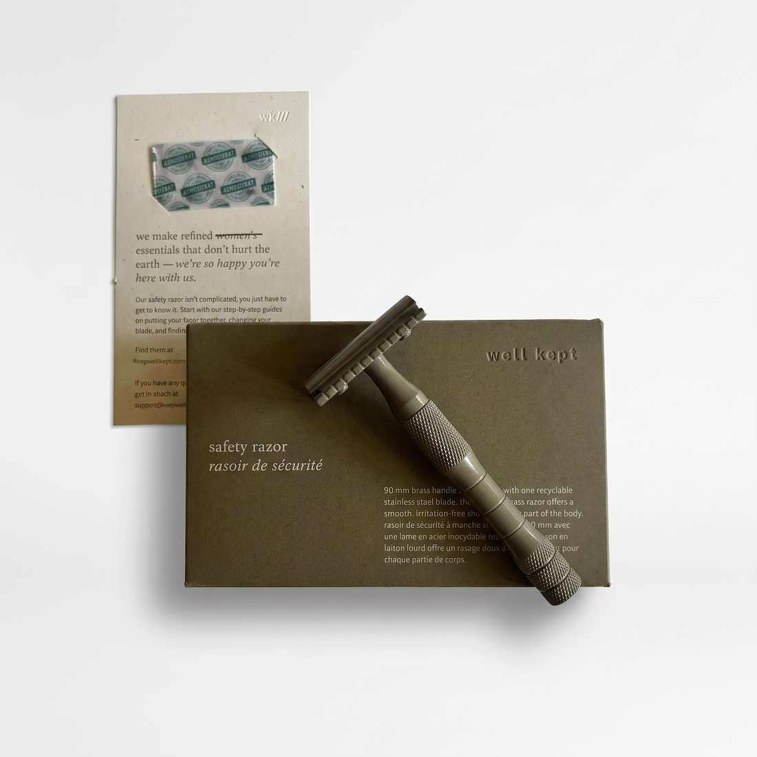 Well Kept Kelp brass safety razor, muted green finish, resting on its box with packaging card, front view on white.